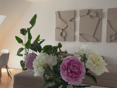 Pan shot of a minimalist, modern living room with the Warrior Series from Armande Darcy, a set of three 3D structural wall art pieces hung on the wall above the beige sofa. A vase with peonies in different shades of pink in the foreground.