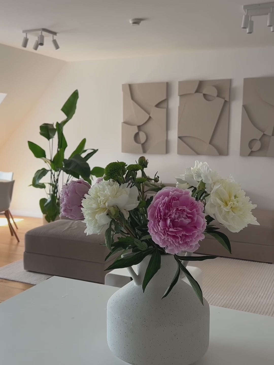 Pan shot of a minimalist, modern living room with the Warrior Series from Armande Darcy, a set of three 3D structural wall art pieces hung on the wall above the beige sofa. A vase with peonies in different shades of pink in the foreground.